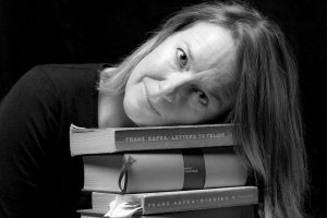 Eine Frau - Autorin Madgaléna Platzová - hat ihren Kopf auf einen Stapel Bücher abgelegt und blickt dabei in die Kamera